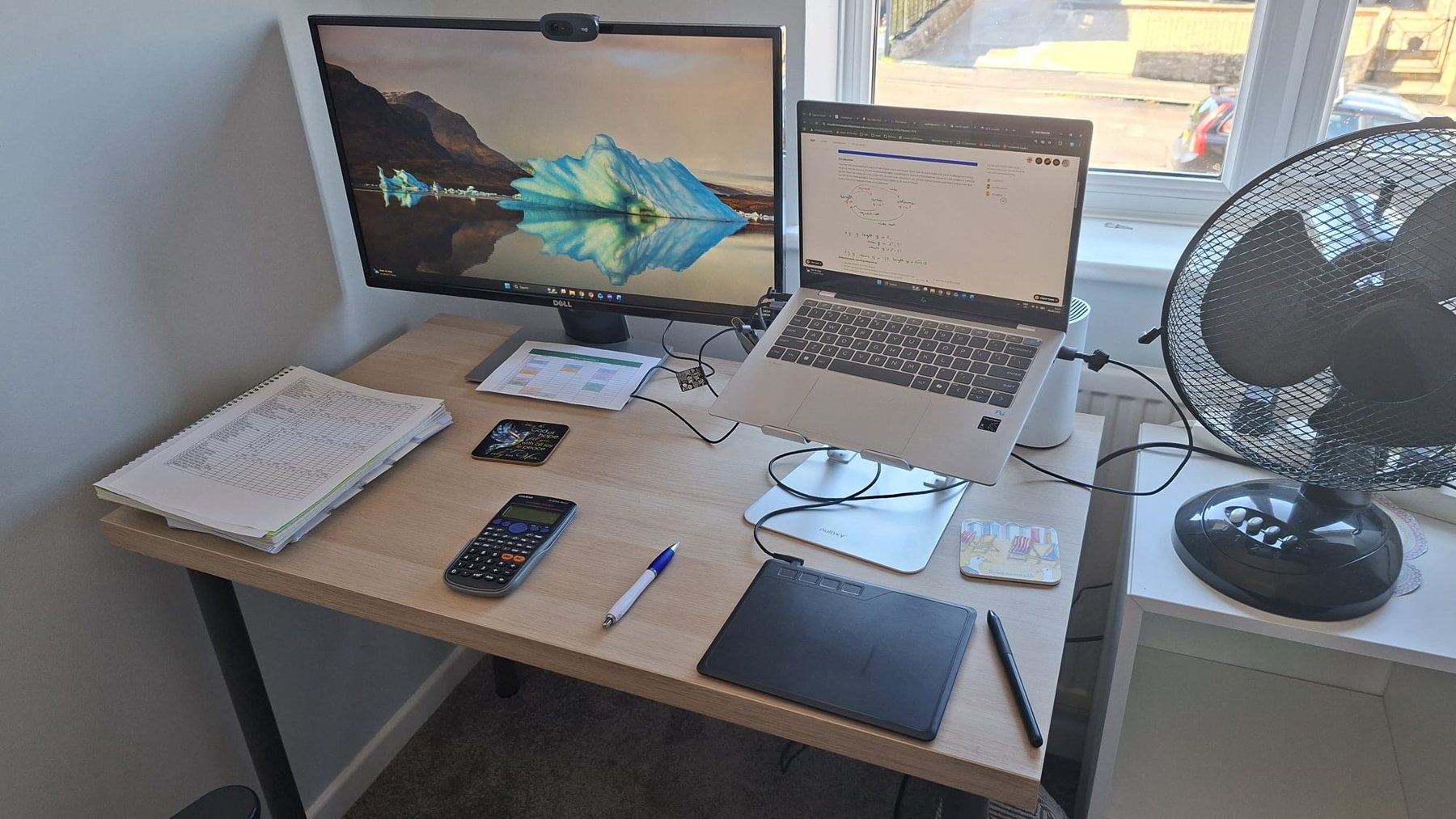 A light wood desk with a notebook, calculator, pen and laptop.