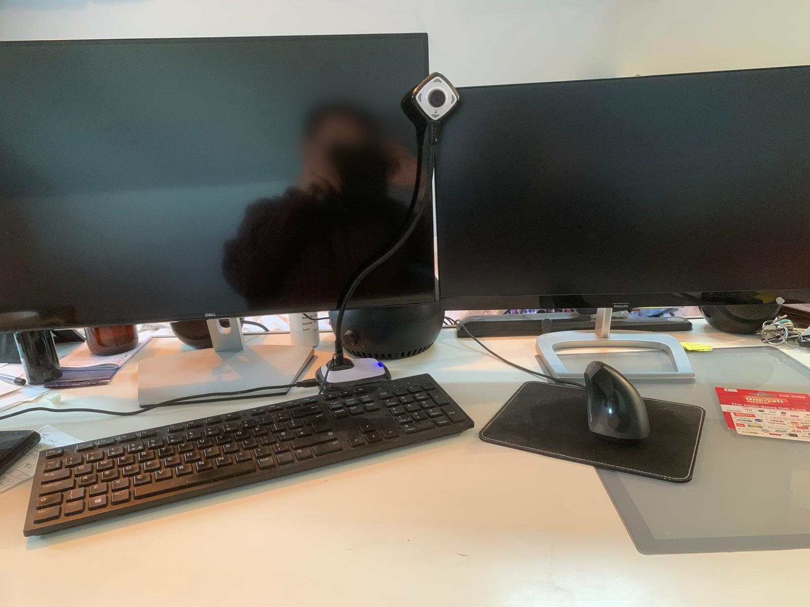 Two monitors, a webcam, a keyboard and a mouse on a pale desk.
