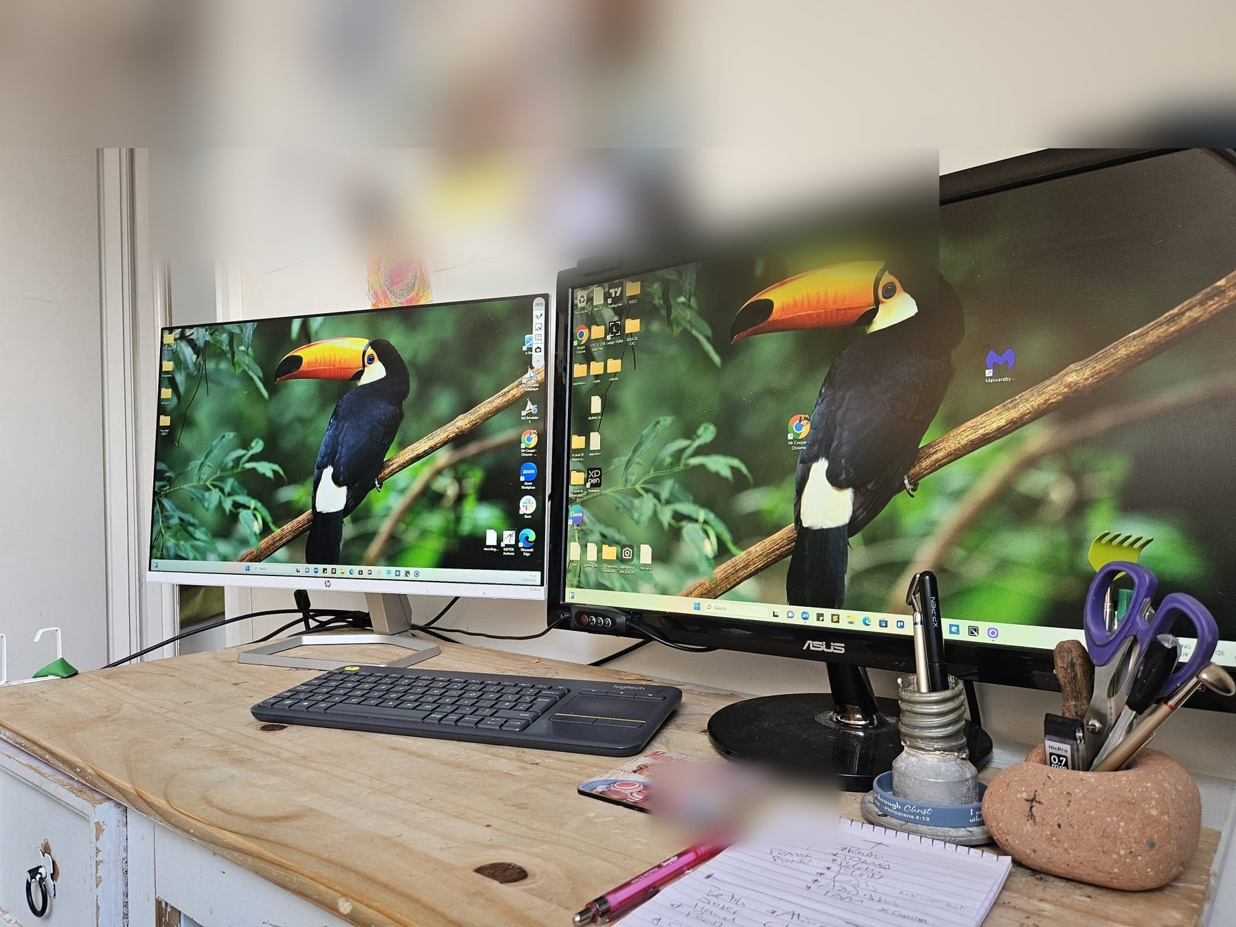 A wooden desk with two monitors, pens and an open notebook.